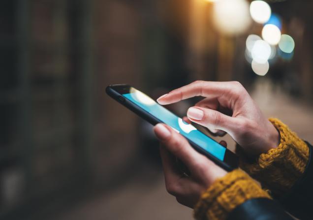 Close up of the hands of a woman using a smartphone. She has brown skin and is wearing a chunky knit ochre jumper. The background is blurred and quite dark. Image by A_B_C via Shutterstock.