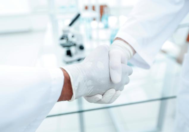 Two people in white labcoats and white rubber gloves are shaking hands. Image by ASDF_MEDIA via Shutterstock