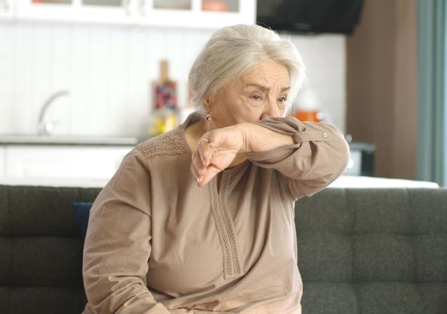 A senior woman coughing into her elbow. She has white skin and long white hair tied back. She's wearing a beige long-sleaved top and sitting on a grey sofa. Image by Red Stock via Shutterstock.