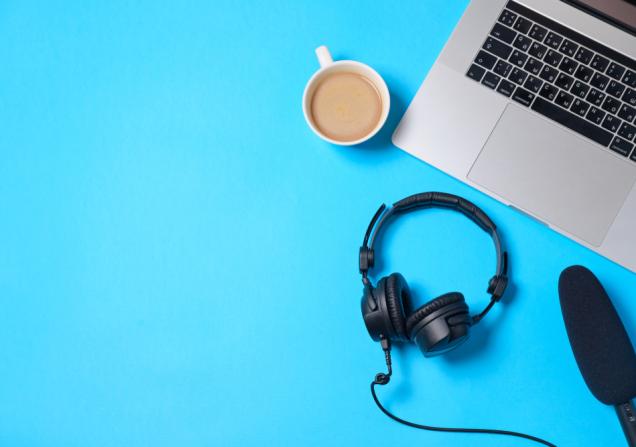 A laptop keyboard, headphones, microphone and a cup of coffee are viewed from above. The background is bright blue. Image by Boiarkina Marina via Shutterstock.