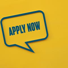 The words 'apply now' written in a speech bubble on a yellow background. Image by Marta Design via Shutterstock.