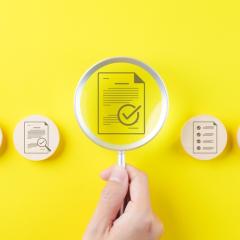 In the centre of the image, a hand is holding a magnifying glass over an icon of a document with a tick mark on it. On either side are more icons with documents and tick marks on wooden circles. The background of the image is bright yellow. Image by SuPatMaN via Shutterstock.