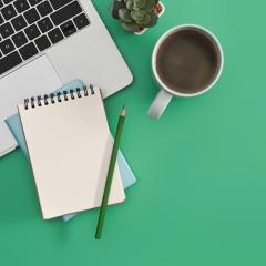 A laptop, spiral-bound notepad, pencil, cup of coffee and a small plant are seen from above. The background is green. Image by Prathankarnpap via Shutterstock.