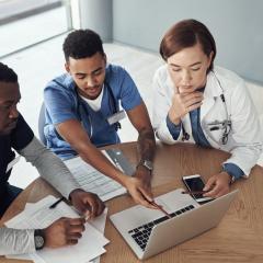 Three people sitting round a table looking at a laptop. Two of them have stethoscopes around their neck. Image by PeopleImages.com - Yuri A via Shutterstock.