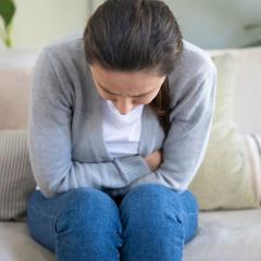 A woman is sitting on a sofa, facing towards us. She's doubled over in pain, so we can't really see her face, just the top of her head. She's wearing a white top, grey cardigan and blue jeans. Her long dark hair is tied back in a ponytail.