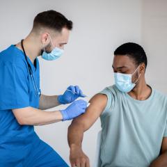 A doctor in blue scrubs gives a man in a pale turquoise t-shirt his COVID-19 vaccination. They're both wearing facemasks. Image by Prostock-studio via Shutterstock