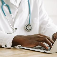 A close-up of a doctor typing at a computer. The doctor is wearing a white doctor's coat and a stethoscope. Image by Ivan Samkov via Pexels.