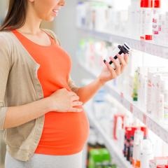 A pregnant woman in an orange t-shirt and beige cardigan looks at the label on a bottle of medicine. Her right hand rests on her bump. Image by Syda Productions via Shutterstock.