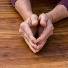 The hands of a man, held lightly together, resting on a wooden table top. The joints of the man's knuckles are visibly swollen. 