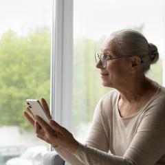 A senior lady using a smartphone. She has long grey hair tied back in a bun and glasses, and is wearing a light coloured sweater. She's sitting in front of a window. Image by fizkes via Shutterstock.