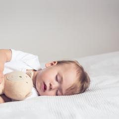 A baby is lying sleeping on its left side on a bed. The baby is cuddling a beige teddy bear.