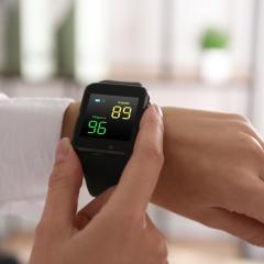 Close-up of a woman measuring oxygen levels with a smartwatch. The watch is on her left arm and she is holding the black watch with the thumb and index finger of her right hand. Image by New Africa via Shutterstock