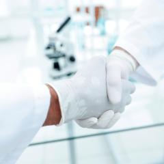 Two people in white labcoats and white rubber gloves are shaking hands. Image by ASDF_MEDIA via Shutterstock
