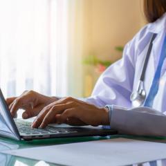A woman doctor wearing a white coat and with a stethoscope around her neck typing at a laptop. Image by NIKCOA via Shutterstock.